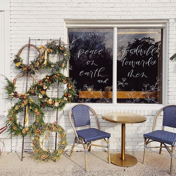 handmade wreaths and cafe chairs in front of a white brick wall and window that reads 'peace on earth, goodwill towards men' in script letters