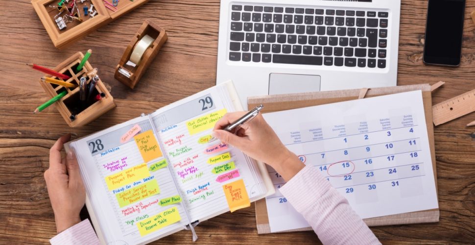 A photo of hands writing something on a planner in front of a laptop with phone, tapes, and pen holder on the table.