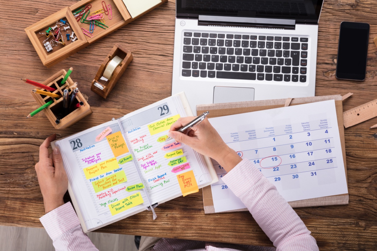 A photo of hands writing something on a planner in front of a laptop with phone, tapes, and pen holder on the table.