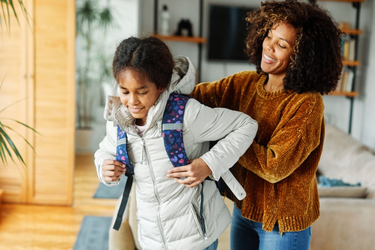 A photo of a mother helping her daughter on her back for school.